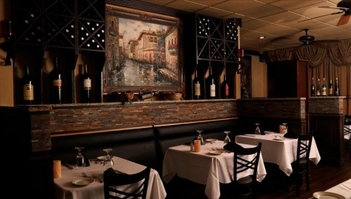 Elegant restaurant interior with white tablecloths, wine bottles on display, and a large painting of a canal scene.