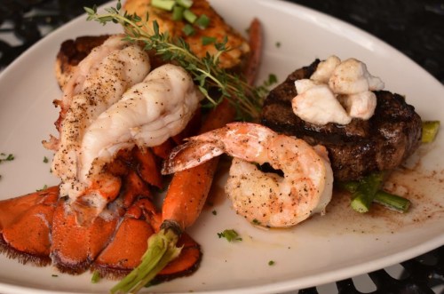A gourmet plate featuring grilled lobster, shrimp, and a beef filet, garnished with herbs and spices.
