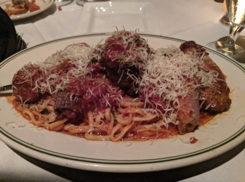 A plate of spaghetti topped with meatballs, sausage, marinara sauce, and grated cheese, served on a white tablecloth.
