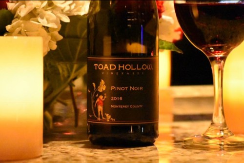 A bottle of Toad Hollow Pinot Noir 2016 on a table with candles and a glass of red wine.