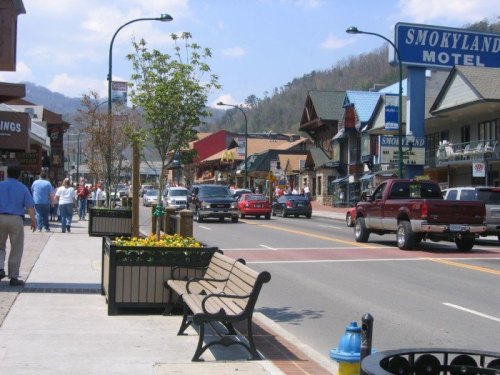 A busy street scene with shops, parked cars, and benches, set against a backdrop of mountains.
