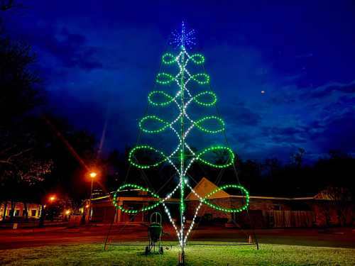 A brightly lit Christmas tree made of green and blue lights against a dark evening sky.