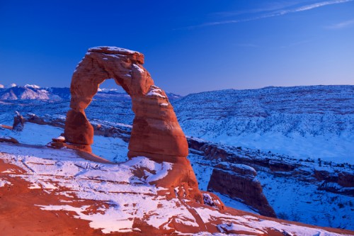 Delicate Arch in Arches National Park, surrounded by snow and mountains under a twilight sky.