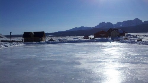 A frozen lake reflects sunlight, surrounded by snow-covered mountains and rustic cabins under a clear blue sky.