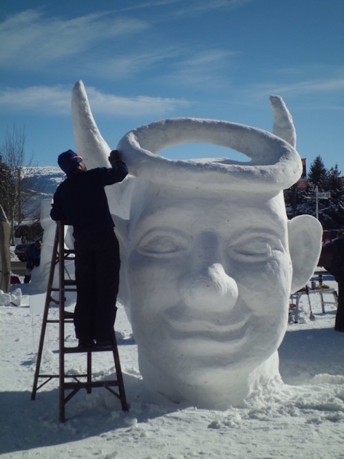 A person carves a large snow sculpture of a smiling face with horns and a halo, standing on a ladder in a snowy landscape.