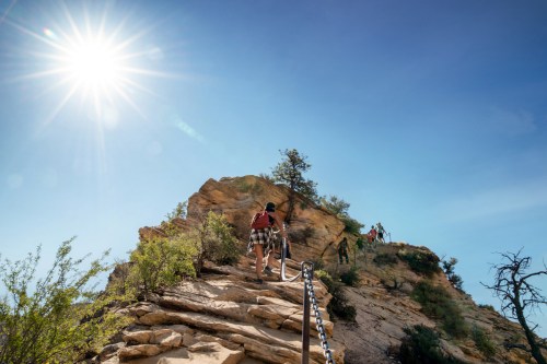 Hikers ascend a rocky trail with a chain, surrounded by desert vegetation and a bright sun overhead.