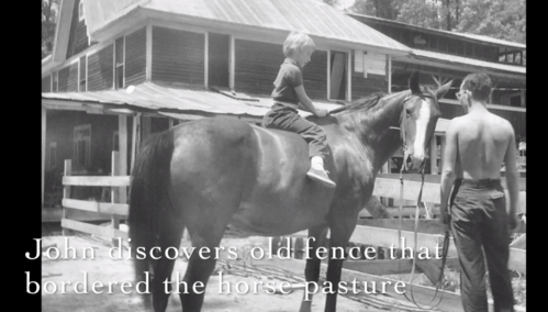 A boy sits on a horse near a barn, while a man stands beside them, with an old fence in the background.