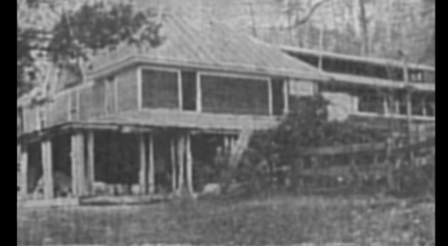 A vintage black-and-white photo of a wooden house on stilts, surrounded by trees and grass.