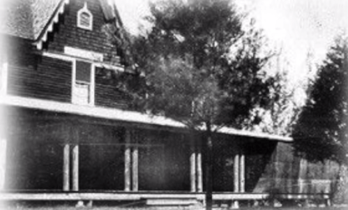 Historic house with a porch, surrounded by trees, featuring a steep roof and wooden siding. Black and white photo.