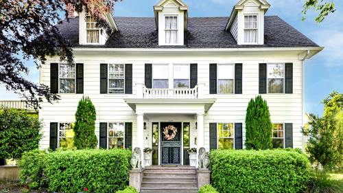 A charming two-story white house with black shutters, a front porch, and lush greenery surrounding it.