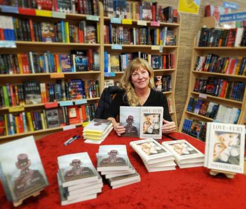 A smiling woman sits at a table with books, surrounded by shelves of colorful books in a bookstore.