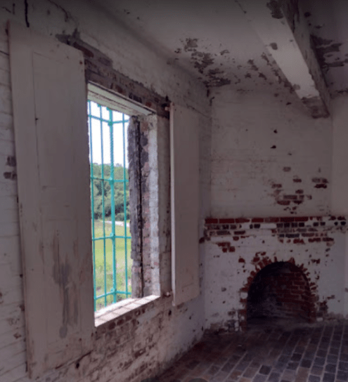 Interior of a weathered room with a window, peeling paint, and a brick fireplace, surrounded by greenery outside.