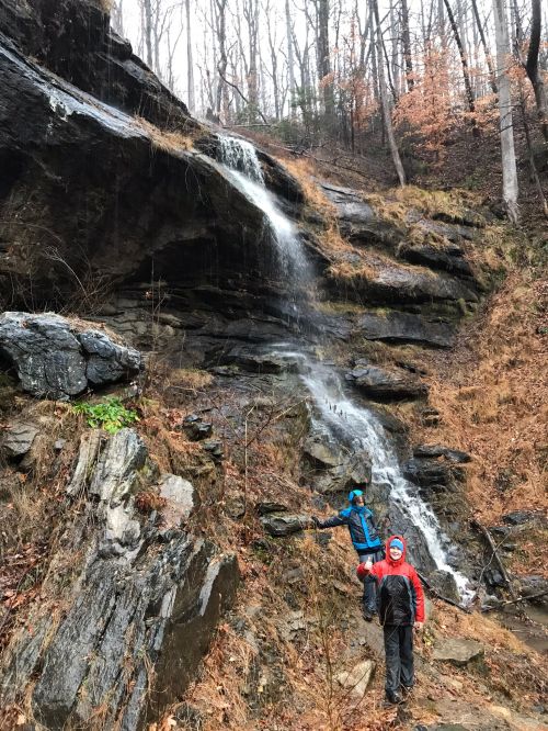 Two hikers stand near a small waterfall cascading down a rocky slope, surrounded by autumn foliage.