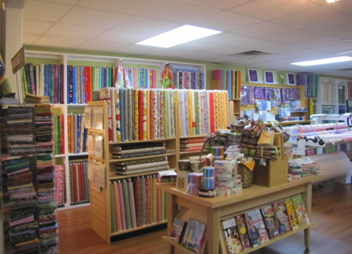 A colorful fabric store interior with shelves of patterned fabrics, a central display table, and quilting supplies.