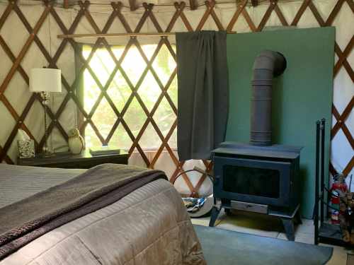 Cozy yurt interior with a bed, wood stove, window, and decorative elements, surrounded by wooden lattice walls.