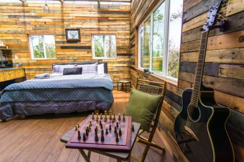 Cozy room with a wooden interior, featuring a bed, a chess set, and a guitar in the corner by large windows.