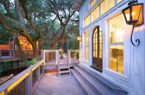 A charming house exterior with large windows, a wooden deck, and lanterns, surrounded by trees at dusk.