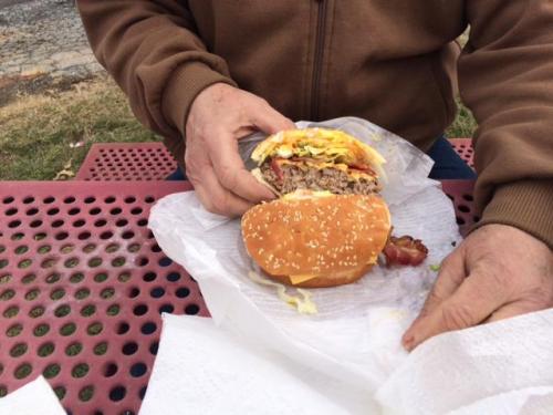A person holding a large burger with layers of meat, cheese, and toppings, sitting at a picnic table.