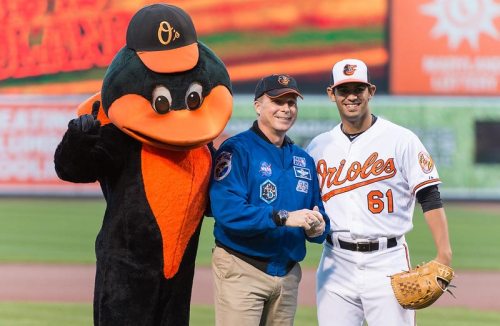 A baseball mascot, an astronaut, and a player in an Orioles uniform pose together on the field.
