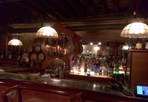 A cozy bar with a wooden counter, illuminated by vintage lamps, showcasing a colorful array of liquor bottles.