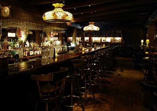 A dimly lit bar with wooden furnishings, vintage lamps, and a long counter lined with various bottles.