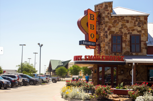A BBQ smokehouse restaurant with a stone exterior and colorful signage, surrounded by parked cars and flowers.