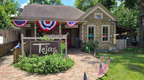A charming wooden building with patriotic decorations, featuring a sign that reads "Tejas" and a welcoming garden.