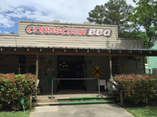 Exterior of Corkscrew BBQ restaurant with a sign, surrounded by greenery and a clear blue sky.