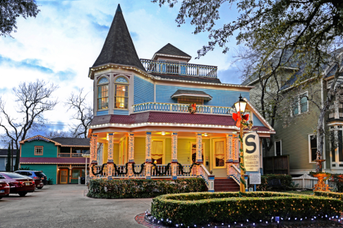 A colorful Victorian-style house adorned with holiday lights, surrounded by trees and parked cars.