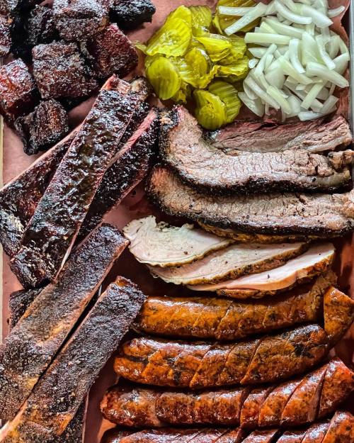 A platter of assorted smoked meats including brisket, ribs, sausage, and pickles, served on butcher paper.