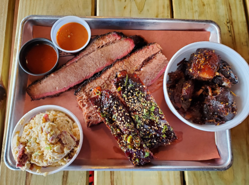A metal tray with sliced brisket, ribs, a side of coleslaw, and two sauces on a pink paper background.