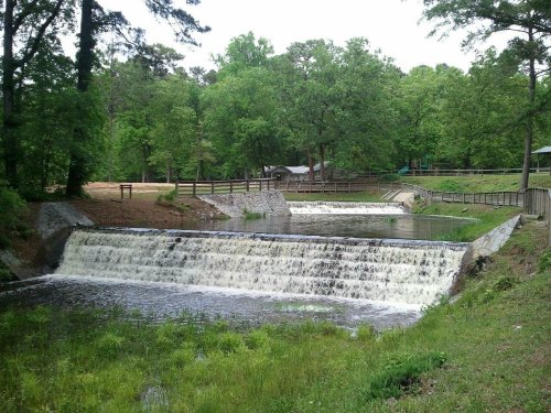 A serene landscape featuring a small waterfall cascading over a dam, surrounded by lush green trees and a peaceful park area.
