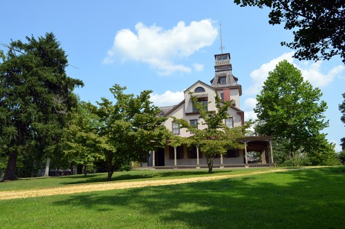 A large, historic house with a tower, surrounded by green trees and a grassy lawn under a blue sky.