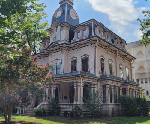 A large, ornate Victorian-style house with a clock tower, surrounded by greenery and blue skies.