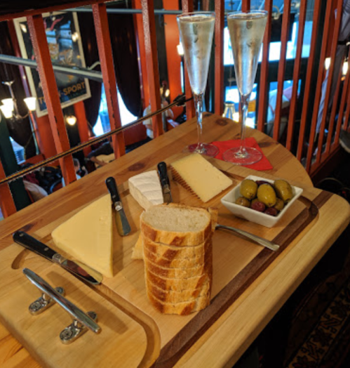A wooden cheese board with various cheeses, bread, olives, and two glasses of champagne in a cozy setting.