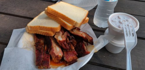 A plate with smoked ribs, two slices of white bread, and two small containers of sauce, next to a bottle of water.