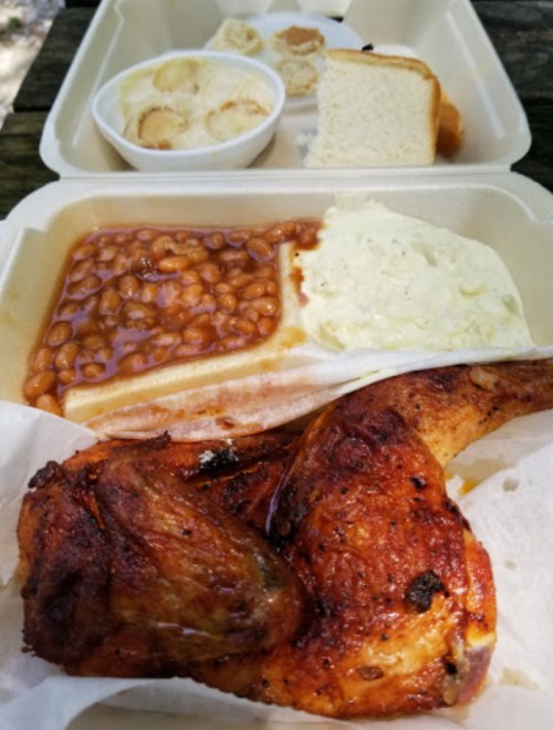 A takeout meal featuring grilled chicken, baked beans, mashed potatoes, bread, and a dessert of banana pudding.