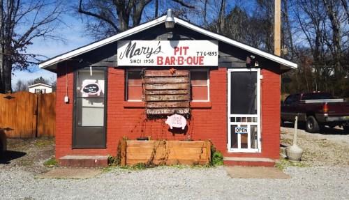 A small red brick building with a sign reading "Mary's Pit Bar-B-Que," featuring an open door and gravel parking.