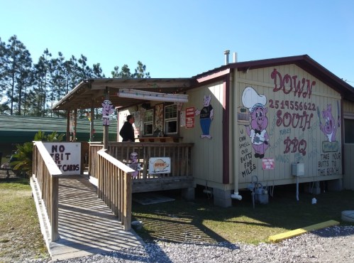 A small BBQ restaurant with a wooden deck, colorful signage, and a person at the counter in a rural setting.