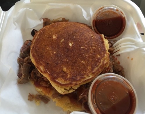 A plate with pulled pork on a cornbread pancake, accompanied by two cups of barbecue sauce.