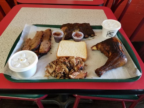 A tray of barbecue featuring sliced brisket, pulled pork, a chicken leg, bread, and two cups of sauce.
