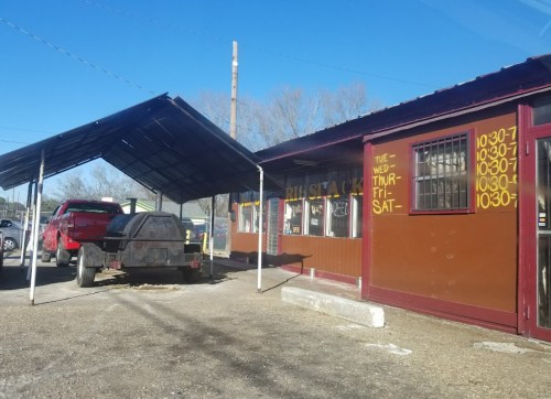A small roadside eatery with a red truck and trailer parked outside, featuring a covered area and business hours painted on the wall.
