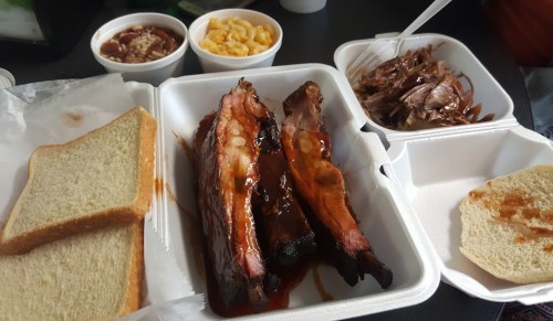 A takeout container with BBQ ribs, pulled pork, macaroni and cheese, baked beans, and slices of bread.