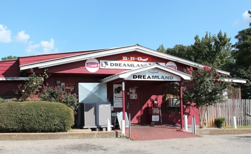 A red building with a sign reading "Dreamland BBQ," featuring outdoor seating and a clear blue sky.