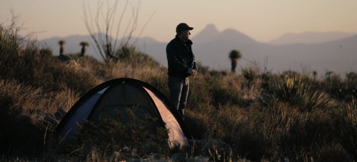 A person stands near a tent in a desert landscape at sunset, with mountains in the background.