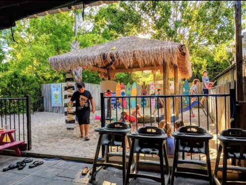 A sandy play area with a thatched gazebo, colorful surfboards, and children playing in a fenced yard.
