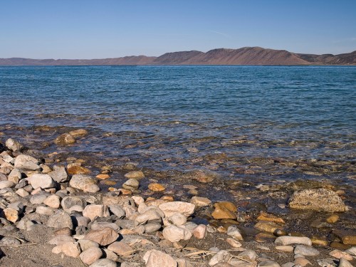 A serene shoreline with smooth stones, clear water, and distant mountains under a blue sky.