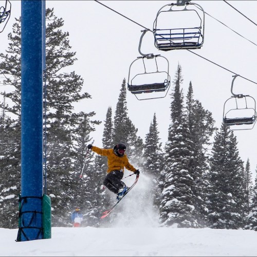 A snowboarder jumps in mid-air near a ski lift, surrounded by snow-covered trees.
