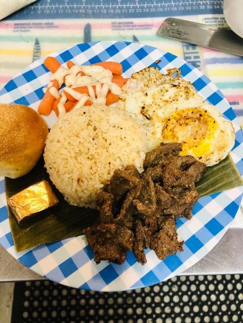 A plate with rice, fried eggs, beef, carrots, a bun, and a small piece of chocolate, arranged on a checkered blue plate.