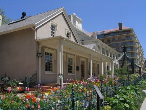 Historic building with a garden of colorful flowers in front, set against a clear blue sky and modern buildings in the background.